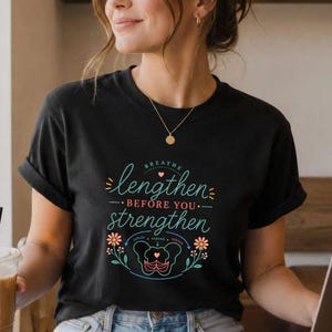 May include: A black short-sleeve t-shirt featuring a graphic print with the text "lengthen BEFORE YOU strengthen" in teal and red, accompanied by a stylized anatomical illustration of a pelvis and floral accents in orange and teal.