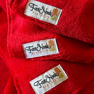May include: Three red fleece blankets stacked on top of each other. Each blanket has a white label with the text "Foot Nook Blanket" in black cursive writing.