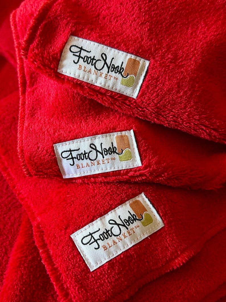 May include: Three red fleece blankets stacked on top of each other. Each blanket has a white label with the text "Foot Nook Blanket" in black cursive writing.