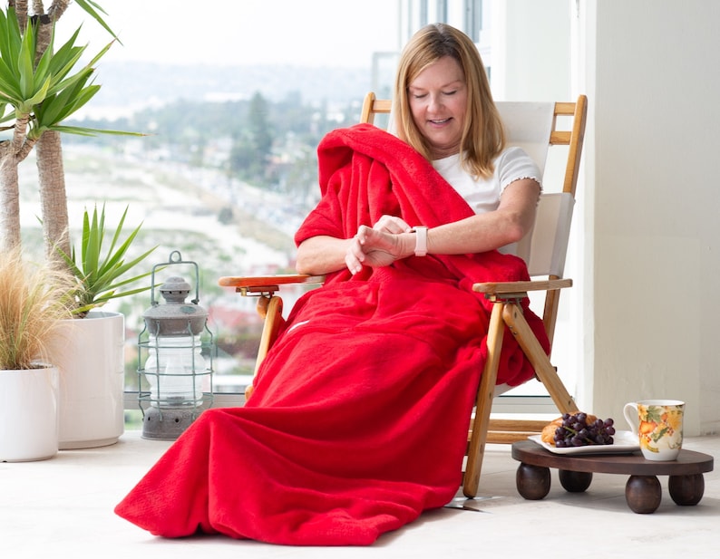 May include: A woman is sitting in a wooden chair, wrapped in a red fleece blanket. She is looking at a smartwatch on her wrist. The chair is on a balcony with a view of the sea.