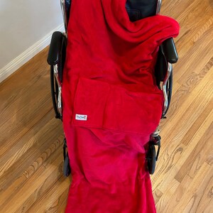 May include: A red fleece wheelchair blanket with a pocket on the front. The blanket is draped over a wheelchair.