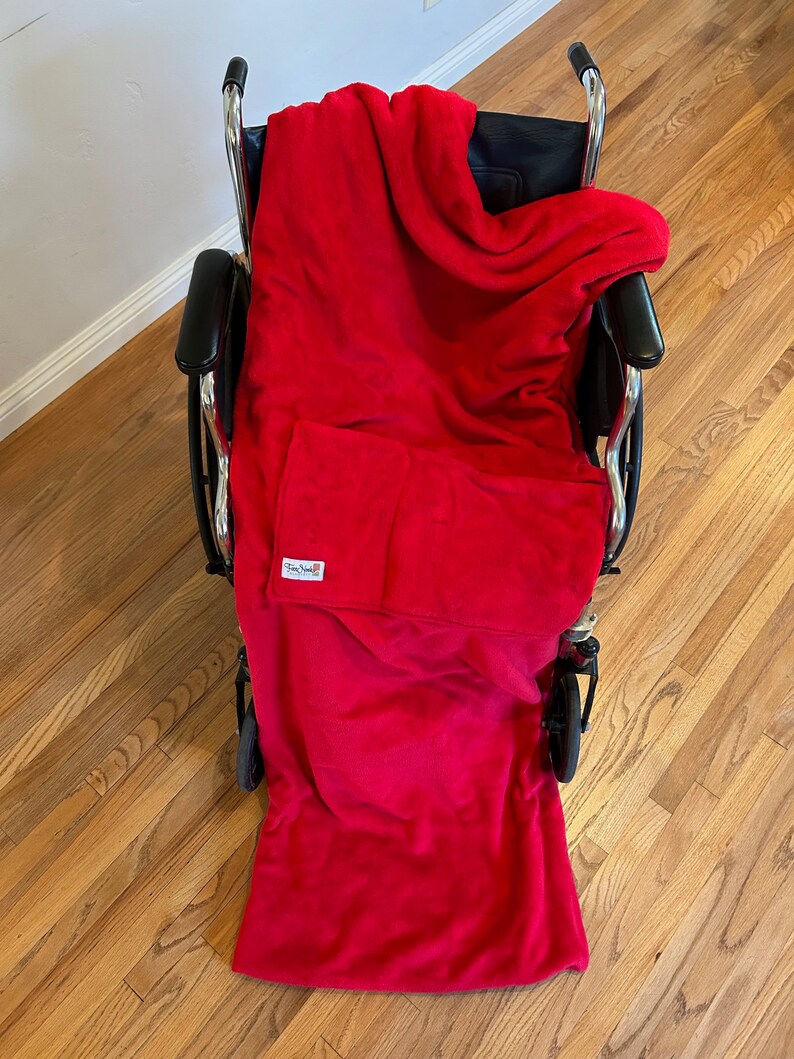May include: A red fleece wheelchair blanket with a pocket on the front. The blanket is draped over a wheelchair.