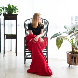 May include: A woman sits in a black wicker chair with a red blanket draped over her legs. She is looking out a window. There are plants in pots on either side of her.