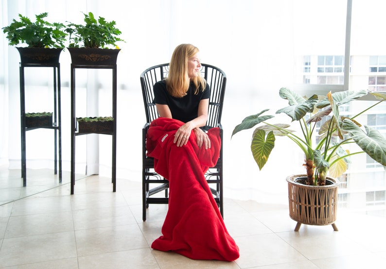 May include: A woman sits in a black wicker chair with a red blanket draped over her legs. She is looking out a window. There are plants in pots on either side of her.