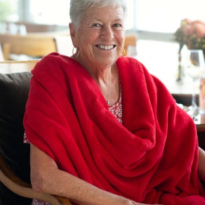 May include: A woman with short, grey hair smiles while sitting in a chair. She is wearing a red blanket draped over her shoulders and a floral print dress.