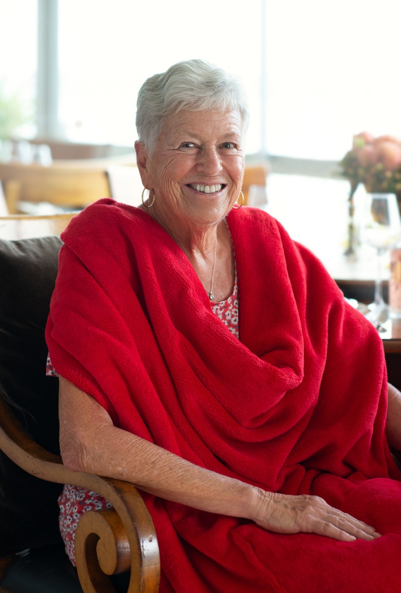 May include: A woman with short, grey hair smiles while sitting in a chair. She is wearing a red blanket draped over her shoulders and a floral print dress.