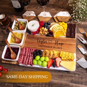 May include: A wooden serving board laden with a selection of cheeses, fruits, and crackers. The board is engraved with the names "Taylor & Hugo" and the date "September 18, 2023". Small white bowls hold dips, and wooden utensils are included.