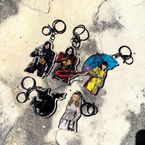 May include: Five acrylic keychains featuring diverse character illustrations: a person holding a baby, a figure in red armour with a guitar, a person in a yellow raincoat under a blue umbrella, a black winged cat, and a person in a white dress.