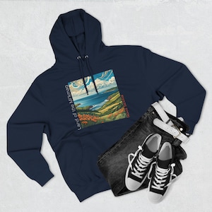 May include: A navy blue hoodie with a graphic print of a landscape scene. The graphic features a coastal view with a blue sky, white clouds, green hills, and a body of water. The text "Land of the Mikmaq" is printed vertically on the left side of the graphic, and the text "Nova Scotia" is printed vertically on the right side of the graphic.