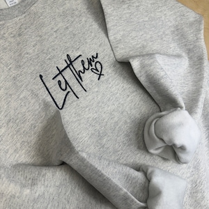May include: A light gray sweatshirt with the words "Let Them" in a handwritten style, with a small heart symbol. The sleeves are cuffed, and the interior is a lighter shade of gray. The sweatshirt is made of a soft material.