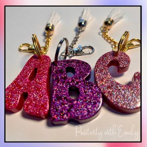 May include: Three resin letter keychains in the shapes of A, B, and C. The A is bright pink, the B is deep purple, and the C is a reddish-pink, all featuring glitter. Each letter has a gold or silver metal keyring, a lobster clasp, and a white tassel.