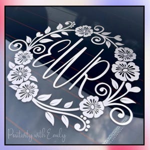 May include: White floral wreath decal with the initials "WR" in the center. The decal is applied to a dark surface, possibly a car window. The text "Positivity with Emily" is at the bottom.