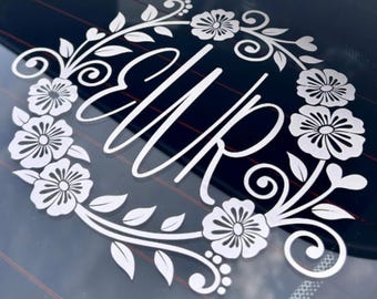 May include: A white vinyl decal featuring the letters "EWR" in a cursive script, centered within a circular wreath design composed of stylized flowers, leaves, and swirling vine accents, applied to a dark, reflective glass surface.