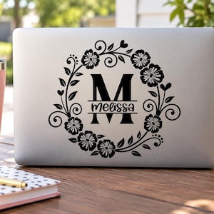 May include: A black vinyl decal applied to a silver laptop lid, featuring a large capital letter M with the name Melissa written in script across the center, all enclosed within a circular wreath of stylized flowers, leaves, and swirling vines.