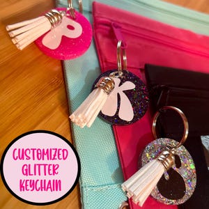 May include: Three glitter keychain charms with white tassels. The charms are pink, purple, and silver with white letters and shapes. The text "CUSTOMIZED GLITTER KEYCHAIN" is visible in the image.
