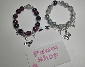 May include: Two beaded bracelets with butterfly charms. One bracelet is red and black, the other is gray. The bracelets are on a white surface with a white card that says "Paw Shop".