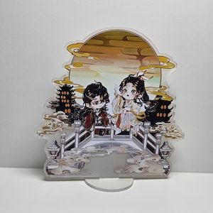 以下が含まれることがあります： 橋の上に立っている2人のアニメキャラクターの印刷された画像がついた白いアクリルスタンド。背景には黄色の月と雲があります。