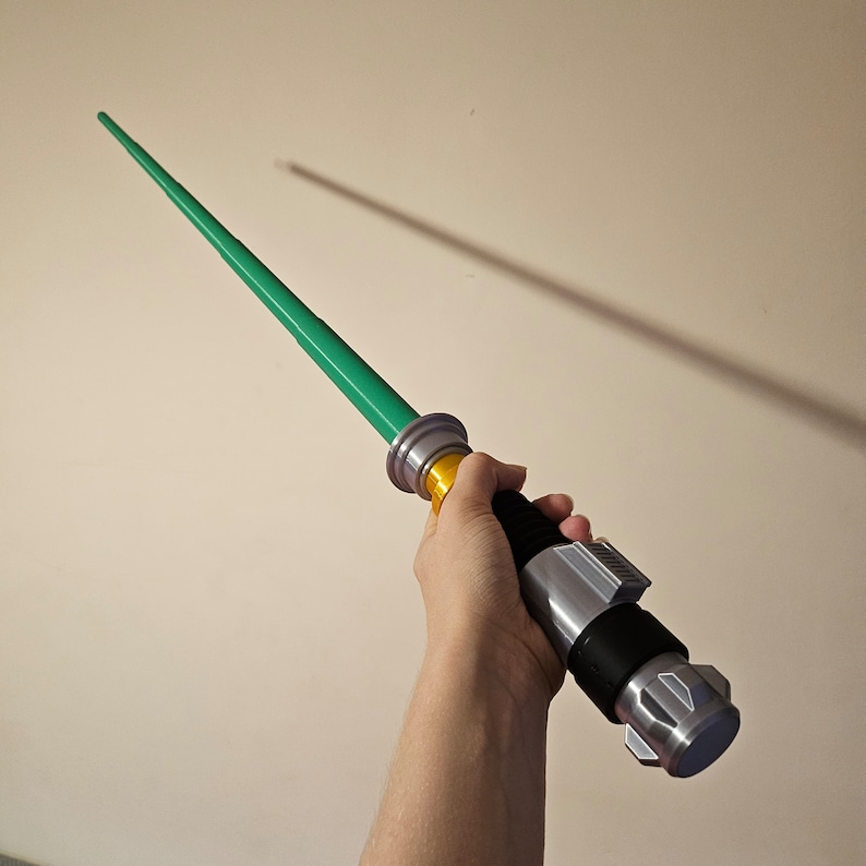 May include: A green lightsaber toy with a silver and gold handle. The lightsaber is held in a person's hand.