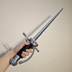 May include: A silver-colored sword with a black handle and a decorative ring. The sword is held by a hand.