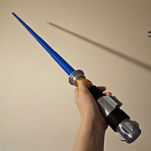 May include: A blue lightsaber toy with a silver and black handle and a yellow ring. The lightsaber is held in a person's hand.