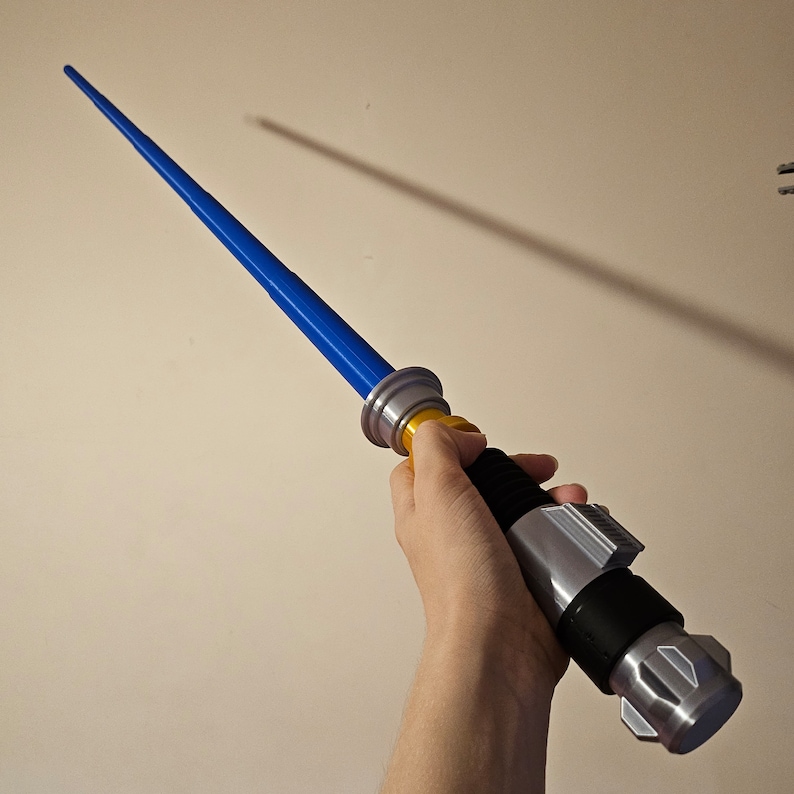 May include: A blue lightsaber toy with a silver and black handle and a yellow ring. The lightsaber is held in a person's hand.