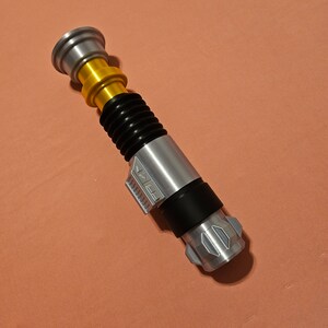 May include: A silver and black lightsaber toy with a gold handle and a black ribbed section. The lightsaber has a silver blade and a black handle with a silver button.
