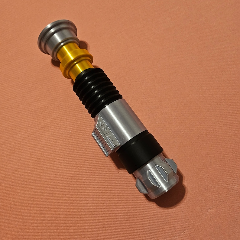 May include: A silver and black lightsaber toy with a gold handle and a black ribbed section. The lightsaber has a silver blade and a black handle with a silver button.