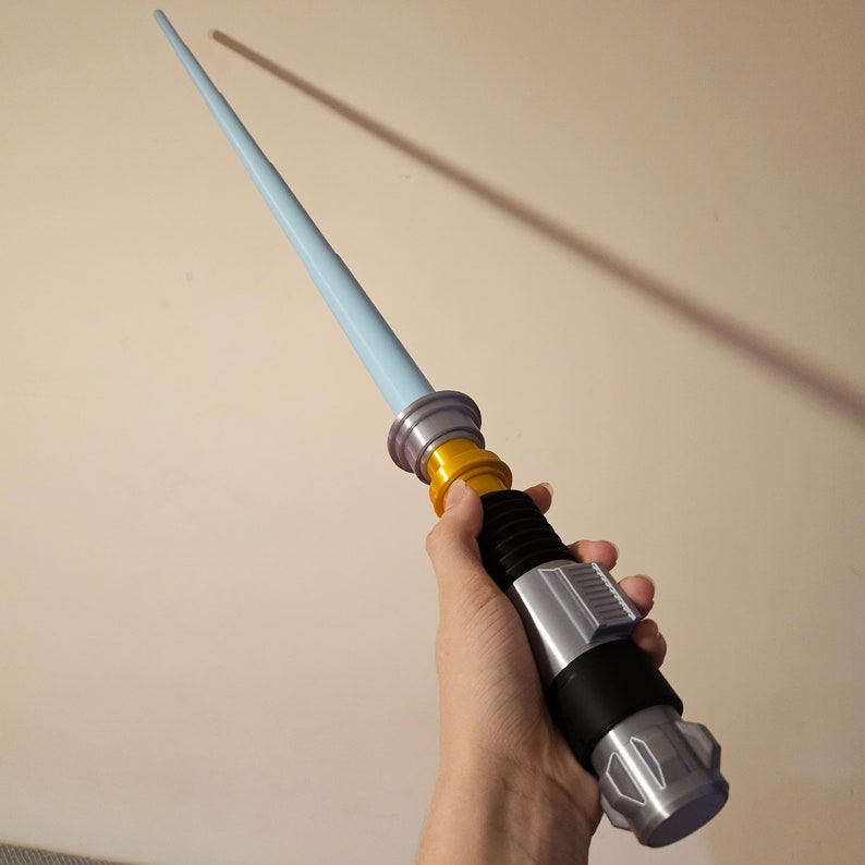 May include: A blue lightsaber toy with a silver and black handle and a gold accent. The lightsaber is held in a hand.