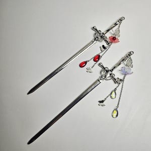 以下が含まれることがあります： 剣を模したデザインの2本のシルバーヘアスティック。それぞれに花と蝶の装飾が施されています。片方は赤い涙型のチャーム、もう片方は黄色と透明のチャームが付いています。白い背景にヘアアクセサリーが配置されています。