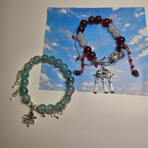 May include: Two bracelets with charms. One bracelet is made of green and white beads with a silver maple leaf charm. The other bracelet is made of red and white beads with a silver lotus flower charm.