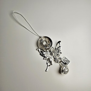 May include: Silver charm featuring a butterfly design with a round charm and a bell. The charm is attached to a white string.