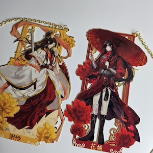 Peut inclure: Deux marque-pages en métal couleur or avec des illustrations de personnages d'anime en rouge et blanc. Les marque-pages présentent des motifs complexes avec des accents floraux et des caractères chinois. Le personnage sur le marque-page de gauche tient une épée et le personnage sur le marque-page de droite tient un parapluie.