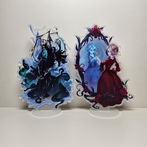 以下が含まれることがあります： アクリル製のキャラクタースタンドフィギュア2体。左側のフィギュアは、王冠をかぶった濃い緑と青の生き物。右側のフィギュアは、青いドレスを着た女性が鏡に映っている鏡の前に立っている赤いドレスを着た女性。両方のフィギュアは、装飾的な花のデザインで囲まれています。