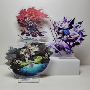 May include: Three acrylic character stand-ups featuring anime characters. The first stand-up features a character with long blonde hair and a blue outfit. The second stand-up features a character with white hair and a blue and purple outfit. The third stand-up features a character with long black hair and a blue and white outfit.