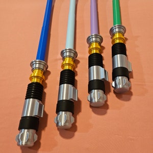May include: Four lightsaber toys with different colored blades: blue, light blue, purple, and green. Each lightsaber has a gold handle and a silver blade. The lightsabers are on a pink surface.