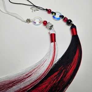 May include: Two decorative tassels with red and white, and black and red silk threads. The tassels are attached to cords with crystal beads, a butterfly charm, and a silver ring.