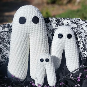 May include: Three white crocheted ghost plushies with black button eyes. The ghosts are arranged in a decreasing size order, with the largest ghost in the back and the smallest ghost in the front. The ghosts are sitting on a black and white floral fabric.