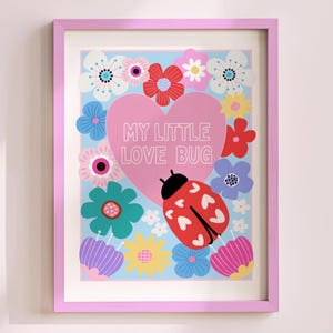May include: A framed art print featuring a pink heart with the text "MY LITTLE LOVE BUG" surrounded by colorful flowers and a red ladybug. The print is in a pink frame and is displayed against a light pink wall.