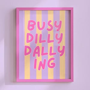 May include: A rectangular wall art print in a pink frame featuring vertical stripes in alternating shades of light purple and pale yellow. The text BUSY DILLY DALLY ING is printed in large, bold, rounded pink letters centered on the striped background.
