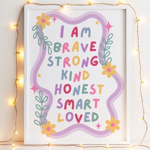 May include: A framed art print with the words "I AM BRAVE STRONG KIND HONEST SMART LOVED" in colorful letters. The design includes a wavy purple border, flowers, and leaves. The frame is white, and the background is a light color.