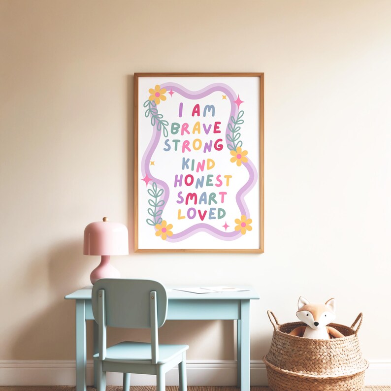May include: A framed print with the words "I AM BRAVE STRONG KIND HONEST SMART LOVED" in colorful text. The print is decorated with flowers and a wavy design. A pink lamp, blue desk, and a fox toy in a basket are in the foreground.