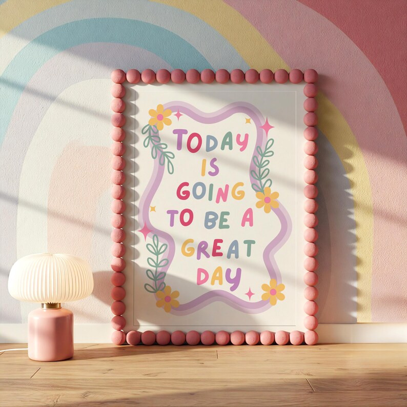 May include: A framed print with the text "TODAY IS GOING TO BE A GREAT DAY" in colorful letters, surrounded by floral and star illustrations. The frame is bordered with pink spheres. A pink lamp with a white shade sits on a wooden surface.