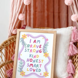 May include: A framed print with the words "I AM BRAVE STRONG KIND HONEST SMART LOVED" in colorful letters. The print is in a light wood frame and is decorated with flowers and leaves. The print is on a wicker chair with pink tassels and pom-poms.