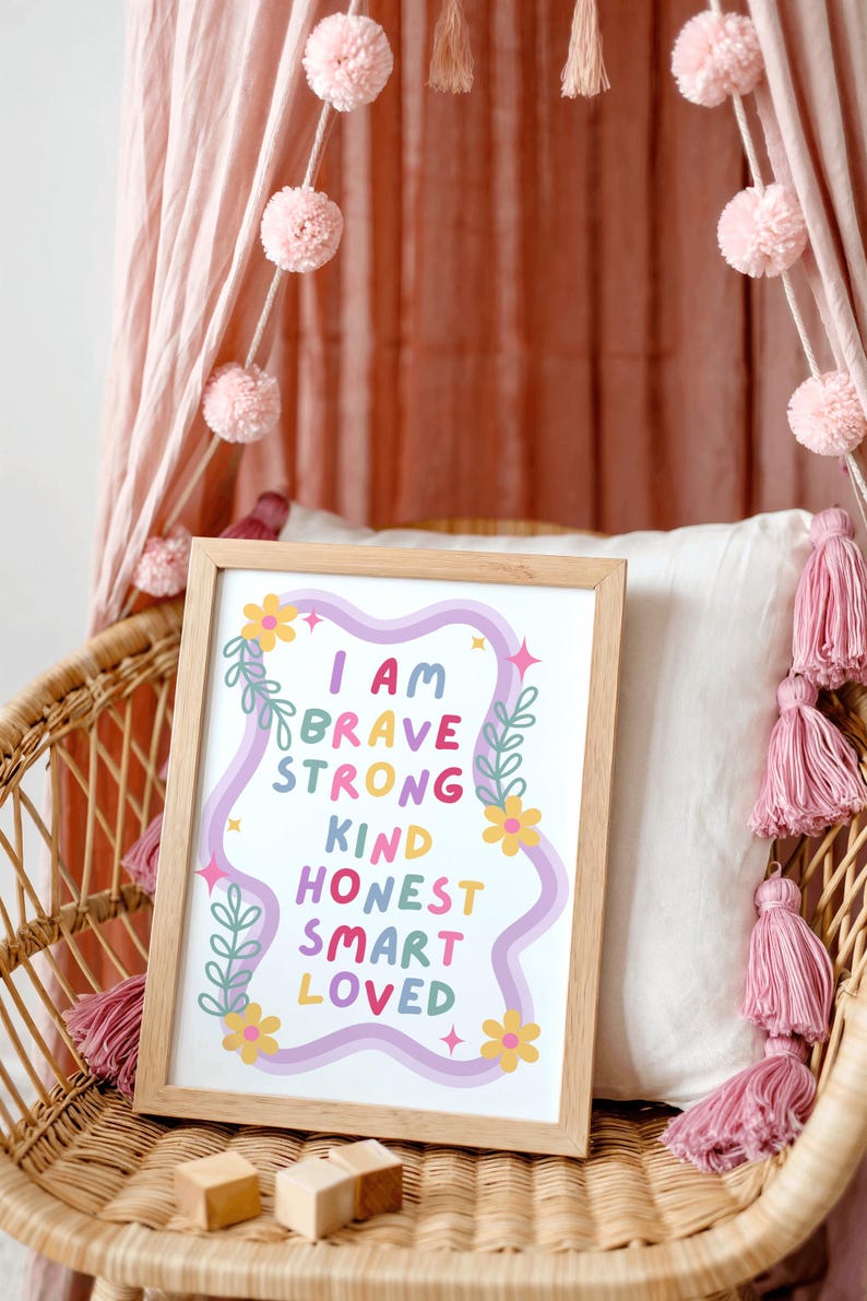 May include: A framed print with the words "I AM BRAVE STRONG KIND HONEST SMART LOVED" in colorful letters. The print is in a light wood frame and is decorated with flowers and leaves. The print is on a wicker chair with pink tassels and pom-poms.