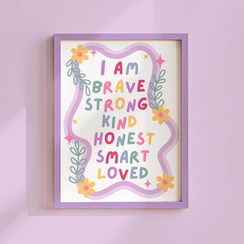 May include: A framed art print with the words "I AM BRAVE STRONG KIND HONEST SMART LOVED" in colorful letters. The print is decorated with flowers, leaves, and stars, and is framed in a lavender frame. The background is a light cream color.