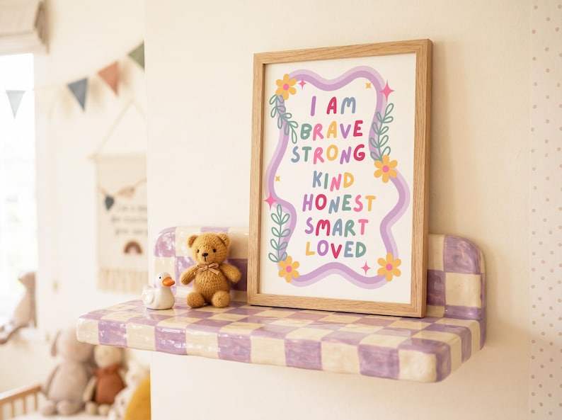 May include: A framed print with the words "I AM BRAVE STRONG KIND HONEST SMART LOVED" in colorful letters. The print is on a shelf with a checkerboard pattern, next to a teddy bear and a duck figurine. The shelf is mounted on a white wall.