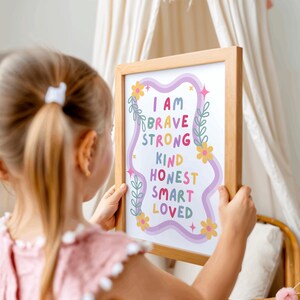 May include: A framed print with the words "I AM BRAVE STRONG KIND HONEST SMART LOVED" in colorful text. The print has a light wood frame and is decorated with floral and star accents. The print is held by a person.