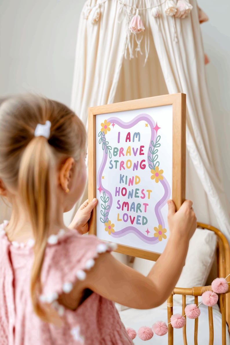 May include: A framed print with the words "I AM BRAVE STRONG KIND HONEST SMART LOVED" in colorful text. The print has a light wood frame and is decorated with floral and star accents. The print is held by a person.
