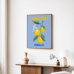 May include: A framed art print featuring a branch of lemons with green leaves and white blossoms against a light blue background. The word "LIMONI" is at the top, and "AMALFI" is at the bottom, both in yellow.
