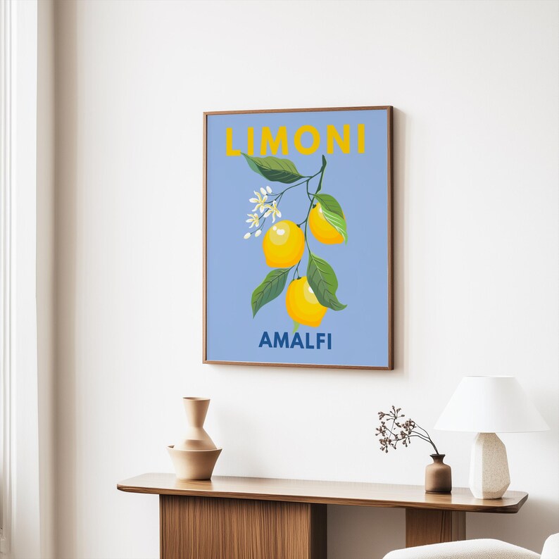 May include: A framed art print featuring a branch of lemons with green leaves and white blossoms against a light blue background. The word "LIMONI" is at the top, and "AMALFI" is at the bottom, both in yellow.